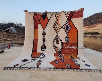 May include: A large, rectangular rug with a cream-colored background and geometric patterns in shades of orange, brown, and blue. The rug has a fringe along the bottom edge and is displayed outdoors.