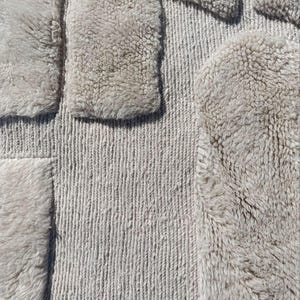 May include: A close-up view of a textured rug in a neutral colour. The rug has a geometric design with a combination of looped and shaggy textures. The soft beige colour would complement a variety of interior design schemes. The rug's dimensions are in centimeters.