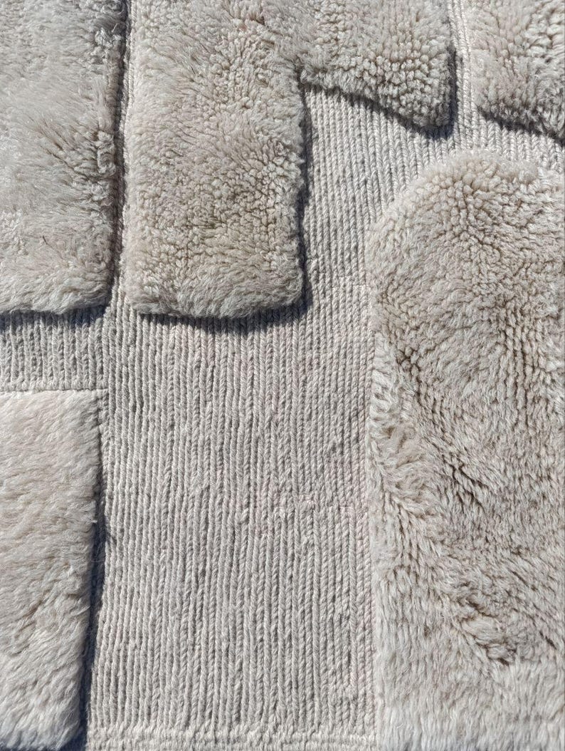 May include: A close-up view of a textured rug in a neutral colour. The rug has a geometric design with a combination of looped and shaggy textures. The soft beige colour would complement a variety of interior design schemes. The rug's dimensions are in centimeters.