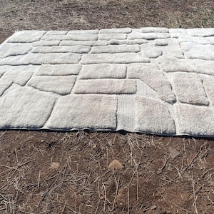 May include: A rectangular, light grey bath mat with a stone-like pattern. The mat is made of a soft, plush material and features a design that resembles individual stone blocks. The mat is placed on a brown surface.