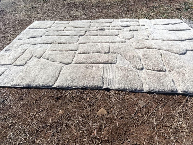 May include: A rectangular, light grey bath mat with a stone-like pattern. The mat is made of a soft, plush material and features a design that resembles individual stone blocks. The mat is placed on a brown surface.