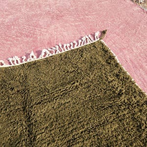 May include: A close-up shot of a textured, olive green rug with a white fringe border. The rug is placed on a pink surface, creating a contrast in colors and textures. The rug's fibers appear soft and plush.
