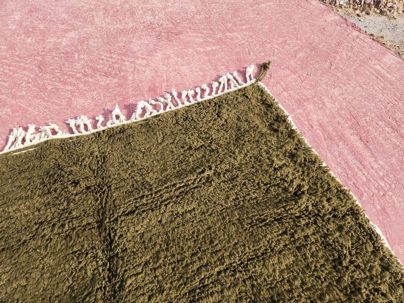 May include: A close-up shot of a textured, olive green rug with a white fringe border. The rug is placed on a pink surface, creating a contrast in colors and textures. The rug's fibers appear soft and plush.