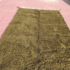 May include: A rectangular olive green rug with a shaggy texture. The rug has a fringe on one side and is displayed on a pink surface. The rug appears to be made of a soft, plush material.