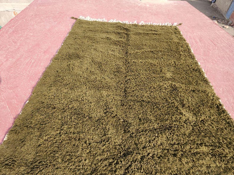 May include: A rectangular olive green rug with a shaggy texture. The rug has a fringe on one side and is displayed on a pink surface. The rug appears to be made of a soft, plush material.