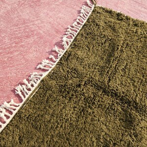 May include: Close-up of a textured, olive green rug with a white fringe border. The rug is partially visible, with a pink surface in the background. The rug's texture appears soft and plush, suggesting a comfortable floor covering.