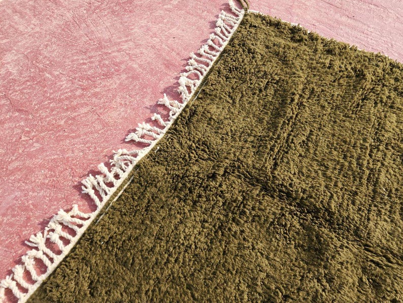May include: Close-up of a textured, olive green rug with a white fringe border. The rug is partially visible, with a pink surface in the background. The rug's texture appears soft and plush, suggesting a comfortable floor covering.