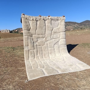 May include: A beige rectangular rug with a geometric pattern is held up against a clear blue sky. The rug has a textured surface with raised rectangular shapes. The top edge has a fringe.