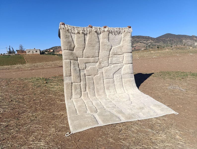 May include: A beige rectangular rug with a geometric pattern is held up against a clear blue sky. The rug has a textured surface with raised rectangular shapes. The top edge has a fringe.