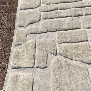 May include: A beige rug with a textured design, featuring various rectangular and irregularly shaped patches. The patches have a fluffy, shaggy appearance, creating a unique, tactile aesthetic. The rug is laid out on a brown surface.
