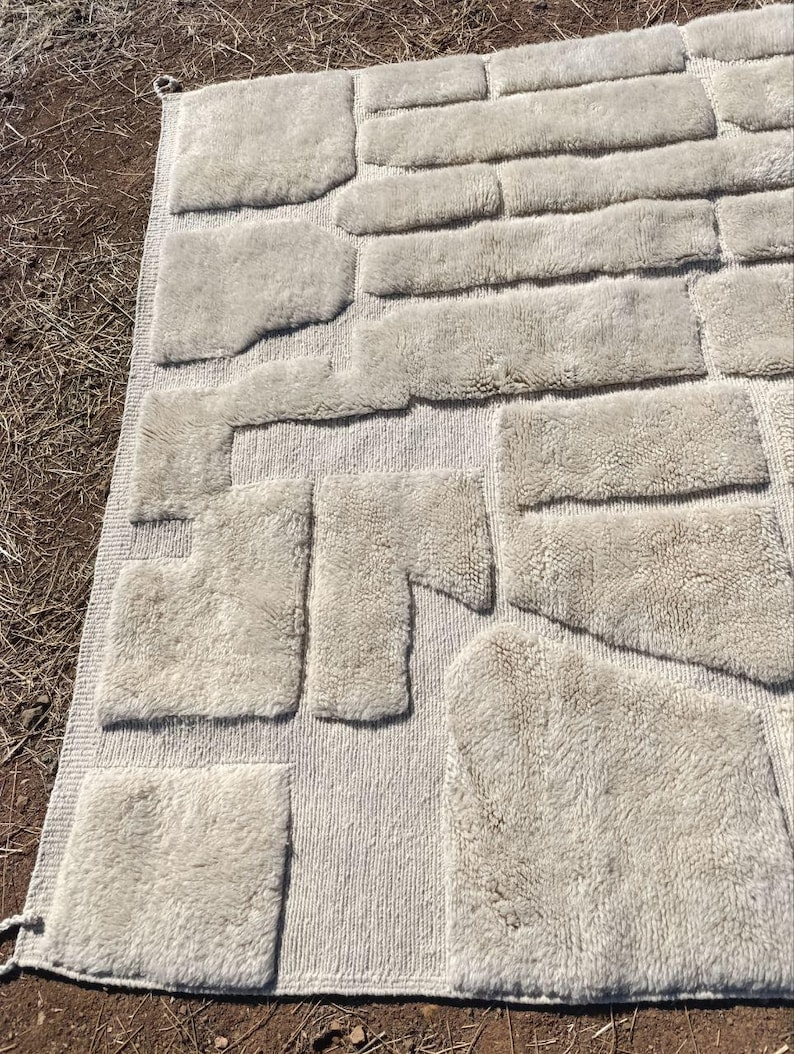 May include: A beige rug with a textured design, featuring various rectangular and irregularly shaped patches. The patches have a fluffy, shaggy appearance, creating a unique, tactile aesthetic. The rug is laid out on a brown surface.