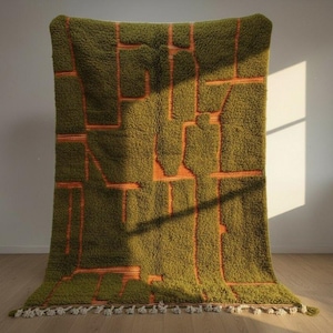May include: A large, olive green rug with a geometric orange design. The rug has a shaggy texture and a fringe of white tassels along the bottom edge. The rug is displayed against a white wall and hardwood floor.