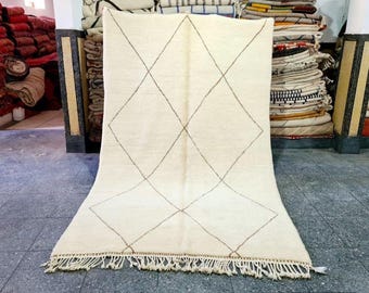 May include: Cream-colored rug with a diamond pattern in brown lines. The rug has a fringe along the bottom edge. The rug is displayed in a room with other rugs and textiles.