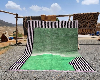 May include: A large, rectangular green rug with a white and green gradient design. The rug features black and white striped borders with purple accents. Other rugs hang in the background.
