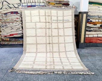 May include: A rectangular, off-white rug with a grid pattern in light brown. The rug has a fringe along the bottom edge and a light brown border. The text "Area-Handmade" is visible on the rug. Stacks of folded textiles are visible in the background.