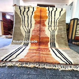 Puede incluir: Una alfombra rectangular grande con un diseño central en tonos naranja y marrón, enmarcada por un fondo gris. Rayas verticales negras y acentos blancos crean un contraste llamativo. La alfombra tiene un borde con flecos y el texto "AreaHandmade".