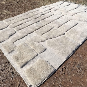 May include: An off-white rectangular rug featuring a raised pattern of rectangular and square shapes. The rug has a fringe on one end. It is displayed outdoors on brown soil and dry grass.