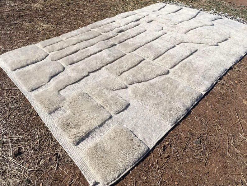 May include: An off-white rectangular rug featuring a raised pattern of rectangular and square shapes. The rug has a fringe on one end. It is displayed outdoors on brown soil and dry grass.