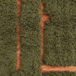 May include: Close-up of a textured rug with a dense, olive green pile. The design features vertical and horizontal stripes in a contrasting orange color, creating a geometric pattern. The rug's texture suggests a soft, comfortable feel.