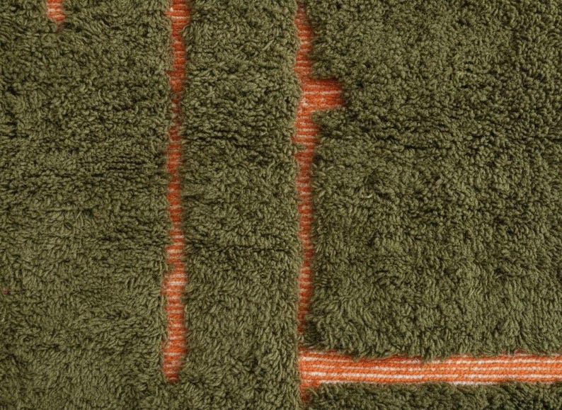 May include: Close-up of a textured rug with a dense, olive green pile. The design features vertical and horizontal stripes in a contrasting orange color, creating a geometric pattern. The rug's texture suggests a soft, comfortable feel.