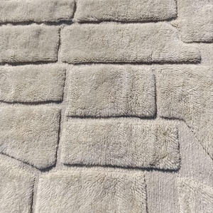 May include: A light grey bath mat with a textured, brick-like design. The rectangular shapes are arranged in a staggered pattern, creating a visual of a stone wall. The mat has a soft, plush appearance.
