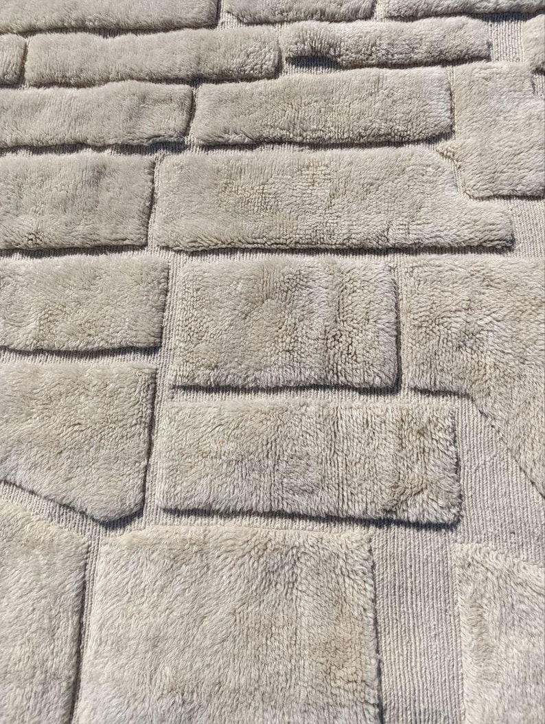 May include: A light grey bath mat with a textured, brick-like design. The rectangular shapes are arranged in a staggered pattern, creating a visual of a stone wall. The mat has a soft, plush appearance.