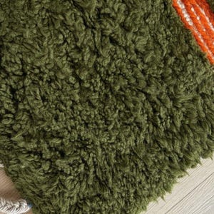 May include: Close-up of a textured, olive green rug with a shaggy pile. A section of the rug features a bold stripe of orange and white. The rug has a braided, off-white border. This rug is suitable for home decor.