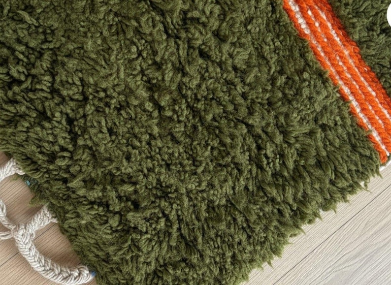 May include: Close-up of a textured, olive green rug with a shaggy pile. A section of the rug features a bold stripe of orange and white. The rug has a braided, off-white border. This rug is suitable for home decor.