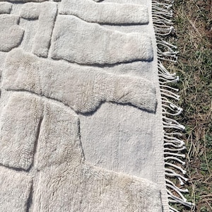 May include: A beige rug with a textured, raised pattern. The design features irregular, rectangular shapes across the surface. The rug has a fringe border on one side, adding a decorative element to the floor covering.