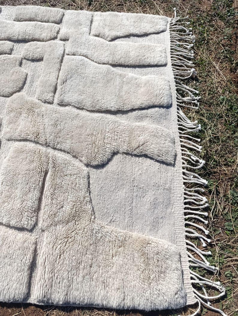 May include: A beige rug with a textured, raised pattern. The design features irregular, rectangular shapes across the surface. The rug has a fringe border on one side, adding a decorative element to the floor covering.
