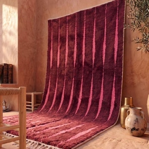 May include: A rectangular area rug featuring a deep burgundy and bright pink vertical striped pattern. The rug has a thick, plush pile texture and is finished with light-colored knotted fringe along the bottom edge, creating a soft, fuzzy appearance.