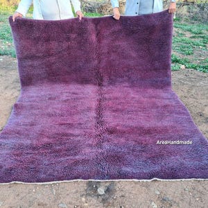 May include: A large, rectangular rug in shades of purple, held up by two people. The rug has a shaggy texture and a gradient effect, transitioning from a darker purple at the top to a lighter shade at the bottom. The text "AresHandmade" is visible.