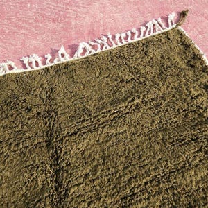 May include: Close-up of a rectangular, olive-green rug with a textured surface. The rug has a white fringe border along one edge. The rug is placed on a pink surface.