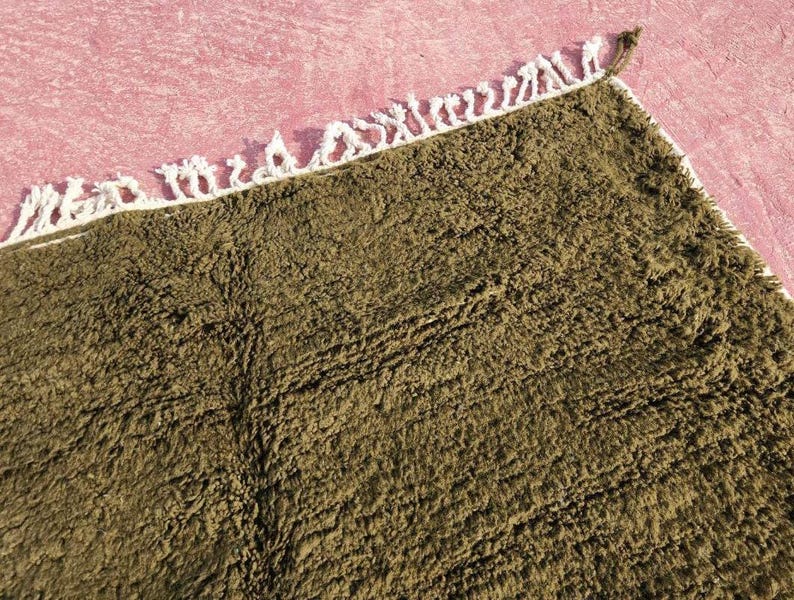 May include: Close-up of a rectangular, olive-green rug with a textured surface. The rug has a white fringe border along one edge. The rug is placed on a pink surface.