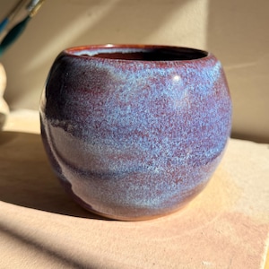 May include: A small, round ceramic bowl with a glossy finish. The bowl is a deep purple with blue and red hues. The interior rim is a darker red. The bowl sits on a light brown surface.