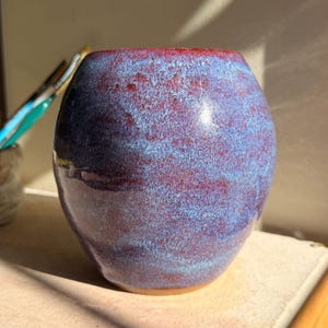 May include: A handmade ceramic vase with a rounded form. The vase displays a mix of blue and red tones, producing a marbled appearance. The rim has a deeper red shade. The vase is placed on a light-coloured surface.