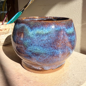May include: A handmade ceramic bowl with a glossy finish. The bowl has a mix of blue, brown, and purple colours. The round bowl sits on a small, unglazed base. A paintbrush rests nearby.