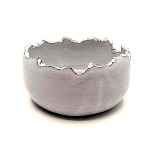 May include: A small, round, grey ceramic bowl with a textured, uneven rim. The rim is highlighted with a white glaze, creating a contrast with the grey interior and exterior. The bowl is likely handmade.
