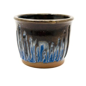 Puede incluir: Una taza de cerámica hecha a mano con un exterior marrón oscuro y un esmalte azul y blanco que gotea por los lados. La taza tiene un diseño texturizado y una base marrón claro.
