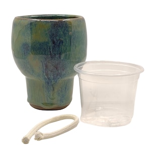 Puede incluir: Una maceta de cerámica con un esmalte verde y azul, un vaso de plástico transparente y una cuerda blanca. La maceta tiene una parte superior ancha y una base más estrecha. La cuerda está enrollada. El vaso está vacío.