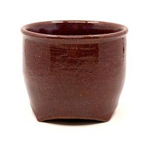 May include: A small, handmade, dark brown ceramic cup with a slightly flared shape. The cup has a smooth, glossy finish and a rounded rim. The interior of the cup is a darker shade of brown. The cup is likely used for drinking.