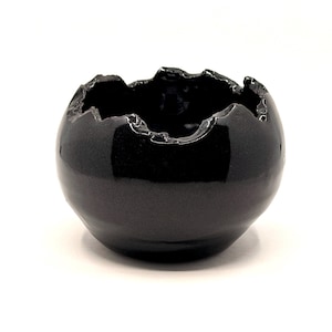 May include: A small, round, black ceramic bowl with a glossy finish. The rim has a jagged, irregular edge. The bowl is set against a plain white background.