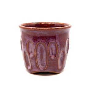 Puede incluir: Una taza de cerámica hecha a mano con un esmalte morado oscuro. La taza presenta una serie de hendiduras ovaladas en el exterior. El borde y el interior de la taza son de un tono morado más oscuro. Una pieza de cerámica única.