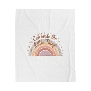 May include: A white blanket with a pastel rainbow design and the text "Celebrate the Little Things". The rainbow features shades of pink, orange, and gray. Decorative elements include clouds, hearts, and stars.