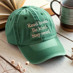 May include: A green baseball cap with the embroidered text "Read books. Be kind. Stay weird." The cap is on a wooden surface next to an open book and a ceramic mug.