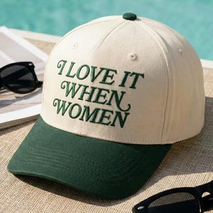 May include: A cream-colored baseball cap with a dark green brim and a dark green button on top. The front of the cap features the embroidered dark green text "I LOVE IT WHEN WOMEN" in a stylized serif font, with a small dark green button on top.