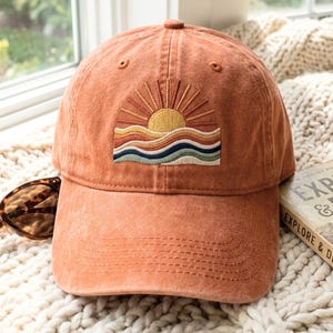 May include: A terracotta-colored baseball cap featuring an embroidered design of a yellow sun rising over multi-colored wavy lines in shades of orange, cream, and blue. The cap has a structured crown, a curved brim, and visible stitching details.