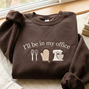 Könnte beinhalten: Ein dunkelbraunes Sweatshirt mit Rundhalsausschnitt und dem gestickten Text "I'll be in my office" in hellem Cremeweiß. Unter dem Text befinden sich gestickte Symbole von zwei Schneebesen, einem gesteppten Ofenhandschuh und einer Küchenmaschine in Creme- und Beigetönen.