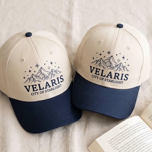 May include: Two baseball caps with a cream-colored crown and a navy blue brim. The caps feature the text "VELARIS CITY OF STARLIGHT" with a mountain and star graphic. The caps are displayed on a neutral background.