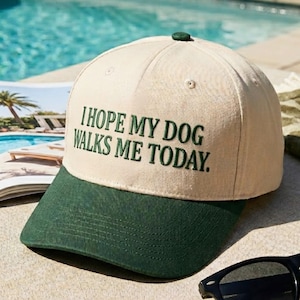May include: A baseball cap with a cream-colored crown and a dark green brim. The cap features the embroidered text "I HOPE MY DOG WALKS ME TODAY." A small green button is on top of the cap.